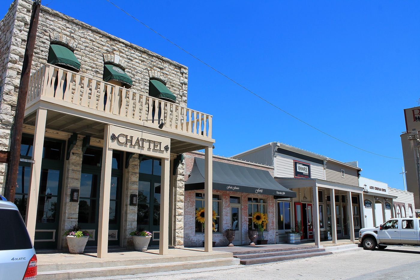 Limestone storefronts with awnings and a second-floor balcony on Front Street in downtown Aledo, Texas, under a clear sky.
